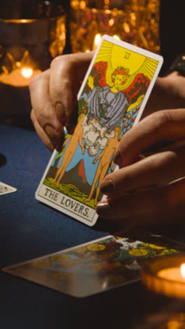 Vertical-Video-Close-Up-Of-Woman-Giving-Tarot-Card-Reading-On-Candlelit-Table-Holding-The-Lovers-Card